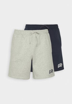 Mężczyzna stoi na białym tle, ubrany w granatowy t-shirt, granatowe szorty ze sznurkiem i logo "GAP", białe skarpetki oraz beżowe sneakersy.