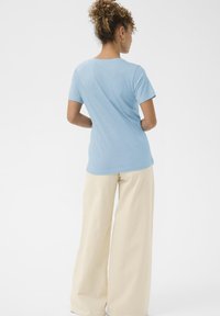 Hellblaues T-Shirt mit kurzen Ärmeln, weicher Stoff, figurbetontes Design, getragen zu cremefarbenen, weit geschnittenen Hosen. Schlichter Rücken, minimale Details.