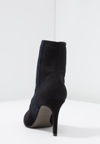 Botín de tobillo negro con un upper de tejido acanalado, franjas laterales en azul en contraste y un elegante tacón de aguja. Fabricado en ante y textil.