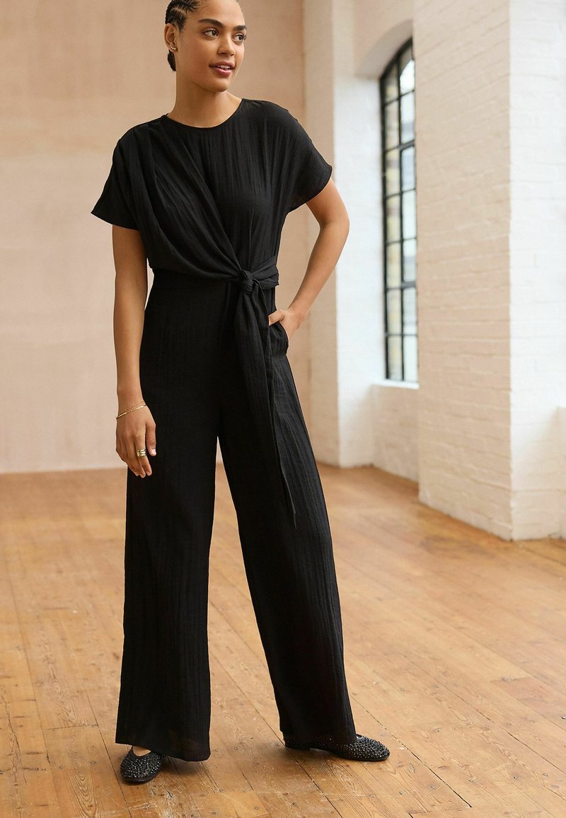 Combinaison noire texturée à manches courtes et détail de taille nouée, avec des jambes larges et une coupe décontractée. Portée avec des ballerines noires ornées.