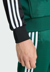 Zeleni sportski outfit s crnim rukavima s manžetama i bijelim prugama, te odgovarajućim zelenim hlačama, ističe teksturu i detalje sportskog dizajna.