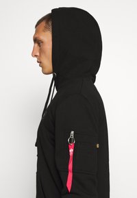 Sudadera con capucha negra que cuenta con un bolsillo con cremallera en la manga con un acento rosa, hecha de un material suave y texturizado.