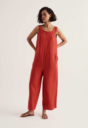 Žena s krátkymi kučeravými vlasmi nosí bezrukávový voľný červený overal a hnedé ploché sandále, stojí s rukami za chrbtom na svetlom pozadí.