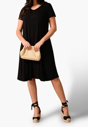 Vrouw draagt een zwarte knielange jurk met korte mouwen, houdt een beige clutch vast en draagt zwarte wedge espadrille sandalen met enkelbandjes.