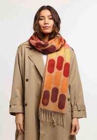 Sciarpa di lana multicolore con forme ovali in rosso, arancione e giallo, con bordi frangiati. La sciarpa è appoggiata su un trench beige.