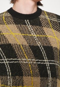 Pletený svetr s černým žebrovaným kulatým výstřihem, hnědým, žlutým a černým kostkovaným vzorem a měkkou texturou.