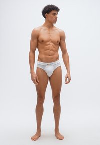 Νεαρός μυώδης άνδρας, όρθιος ξυπόλητος, φορώντας λευκά εσώρουχα με έγχρωμη ελαστική ζώνη με λογότυπο, κοιτάει προς τα αριστερά του σε απλό φόντο.