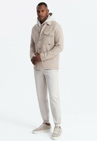 Veste en mélange de laine beige avec fermeture à boutons et poche poitrine, portée sur un sweat à capuche gris, associée à un pantalon de jogging gris clair et des baskets neutres.