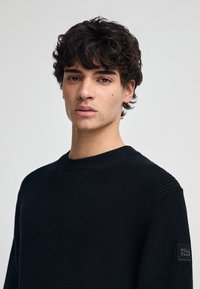 Zwarte gebreide trui met een ronde hals, met ribbelstructuur en een klein logo-label op de linker mouw. Casual pasvorm, minimalistisch design.