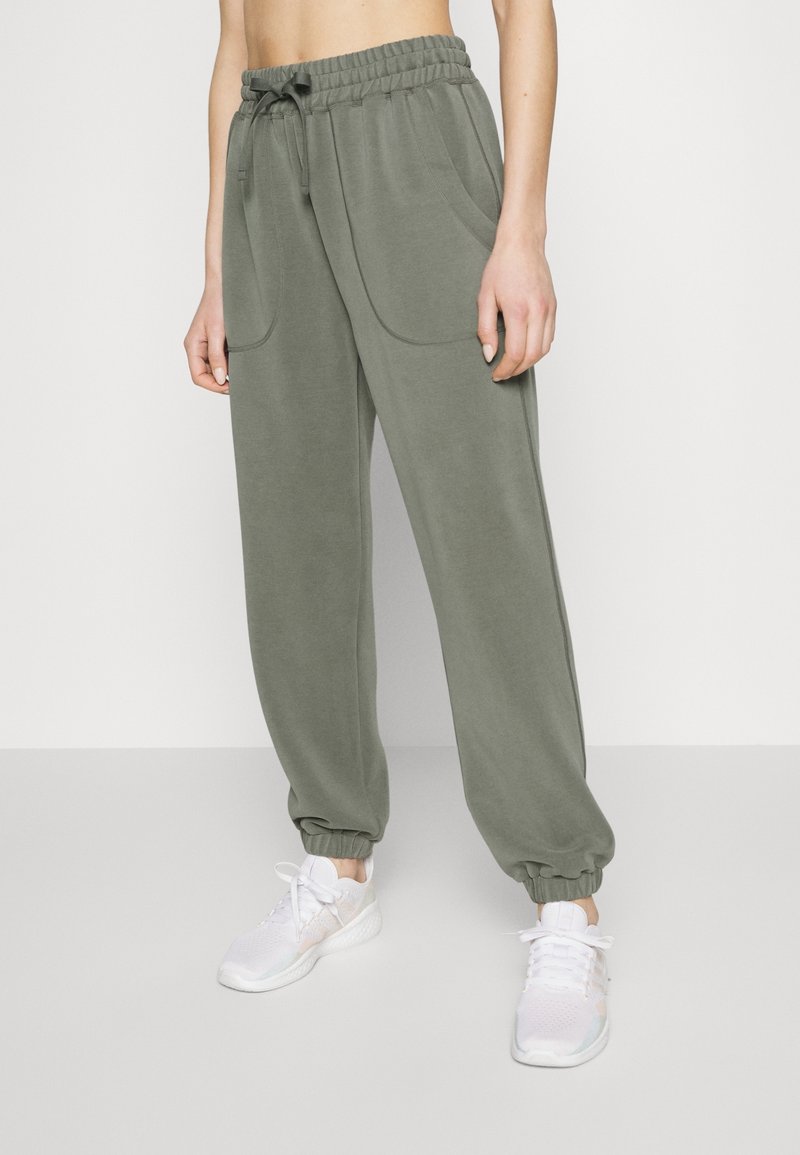 Pantaloni sportivi verde oliva con vita elastica e coulisse, dotati di tasche laterali e polsini sagomati. Indossati con sneakers bianche.