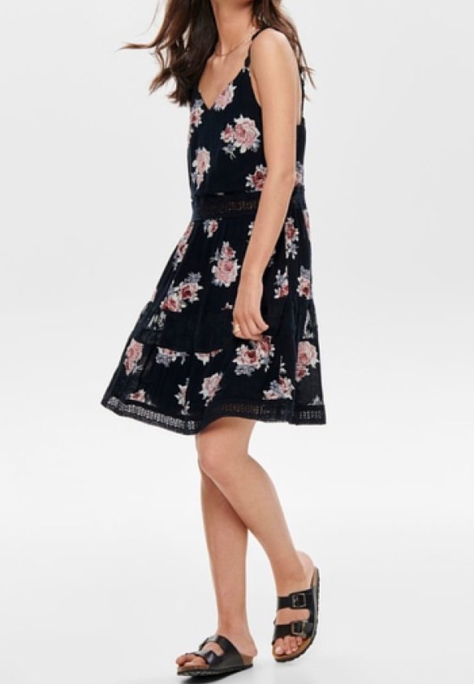 Robe noire imprimée de fleurs avec des bretelles réglables, un design à volants et une bordure en dentelle. Associée à des sandales noires avec une double bride.