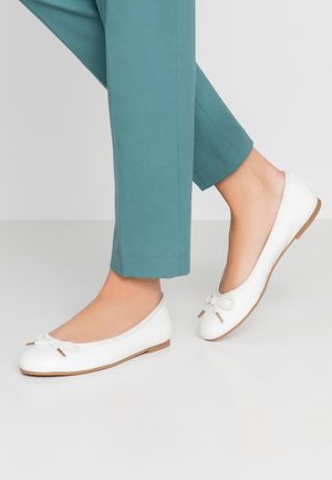 Fødder iført hvide balletsko med sløjfer, kombineret med tealfarvede cropped bukser, mod en ensartet lys baggrund.
