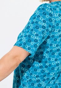 Top a maniche corte in teal con un motivo floreale. Il tessuto presenta diverse tonalità di blu e fiori bianchi su uno sfondo solido.