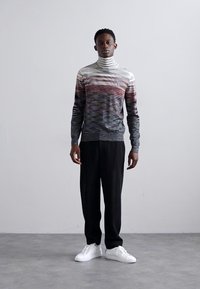 Turtlenecko tipo megztinis su horizontaliomis gradientinėmis juostomis pilkos, raudonos ir grietinėlės spalvų atspalviais. Derinamas su juodomis kelnėmis ir baltais sportbačiais.