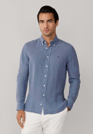 Homme portant une chemise bleu clair boutonnée à manches longues avec un pantalon blanc, debout devant un fond gris clair uni.