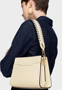 Osoba z krótkimi brązowymi włosami, w ciemnej dżinsowej kurtce, trzymająca kremową skórzaną torbę na ramię z szerokim przyszytym paskiem i złotymi akcentami.