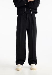 Personne portant un pantalon large noir, une veste noire avec fermeture éclair et des baskets blanches se tenant devant un fond blanc.