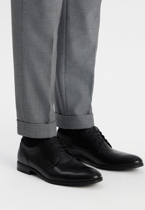 Zapatos de cuero negro con cordones, de textura suave y punta redonda, combinados con pantalones ajustados de un gris claro que tienen puños en el dobladillo.