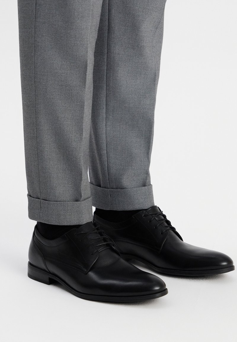 Zwarte leren veterschoenen met een gladde textuur en een ronde neus, gecombineerd met lichtgrijze, op maat gemaakte broeken die omgeslagen zijn aan de onderkant.