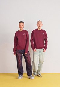 Bordo sporta krekli ar logo uz krūtīm, apvienoti ar tumšām un gaišām vaļīgām biksēm. Modeļi stāv uz dzeltenas grīdas ar vienkāršu fona.