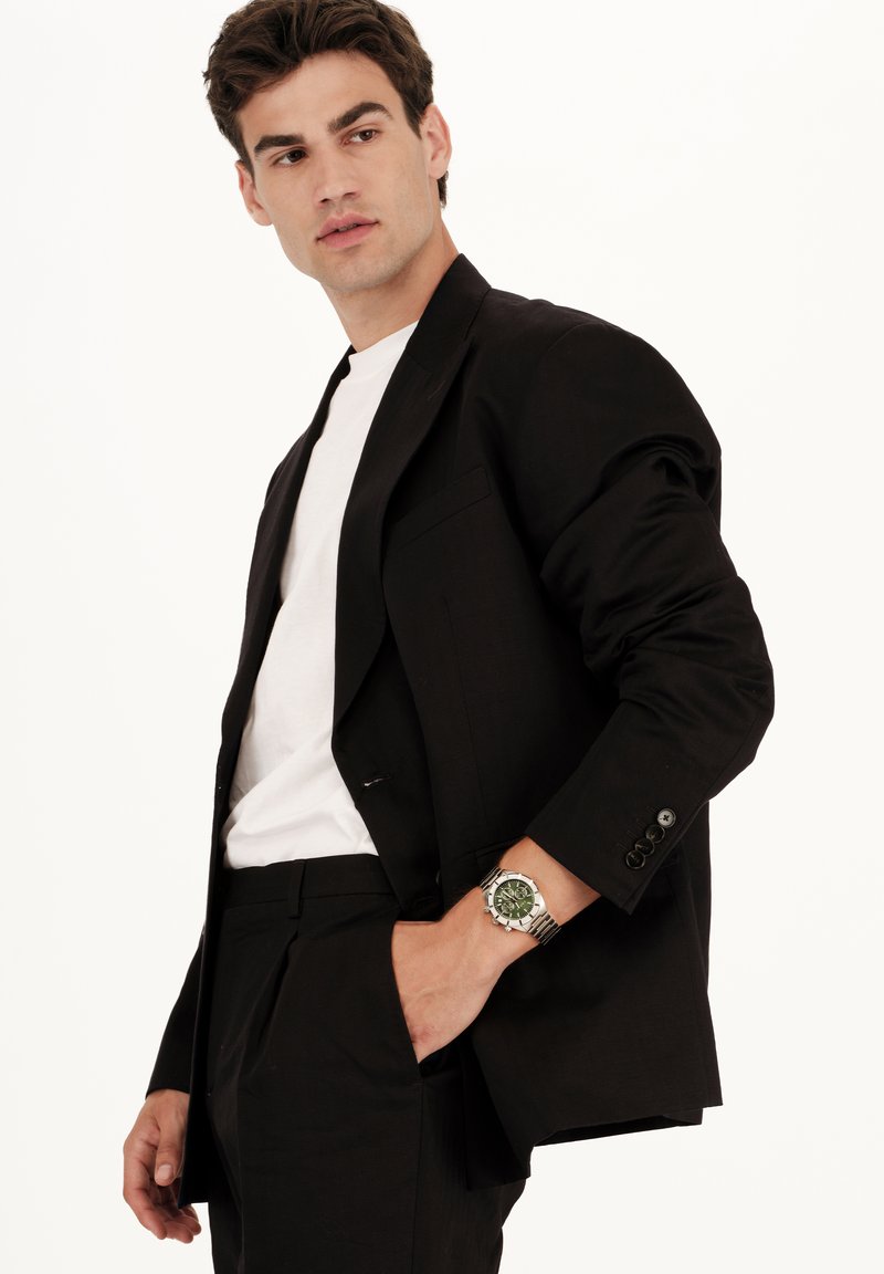 Blazer noir avec une poche unique, porté avec une chemise blanche. Porte une montre argentée au cadran vert, sur un fond clair.