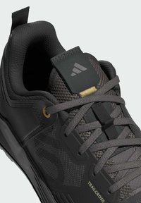 Sort atletiksko i mesh- og syntetiske materialer med tekstureret overflade, grå snørebånd, gulddetaljer og en mærket tungeflap.