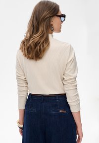 Vrouw met golvend bruin haar, gekleed in een geblokte crèmekleurige lange mouw top, donkerblauwe jeans, schildpadbril en grote ronde oorbellen, met haar rug naar de camera gekeerd.