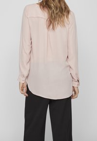 Camicia rosa chiaro a maniche lunghe con orlo arrotondato, realizzata in materiale morbido. Presenta polsini con bottoni e una sezione inferiore leggermente trasparente.