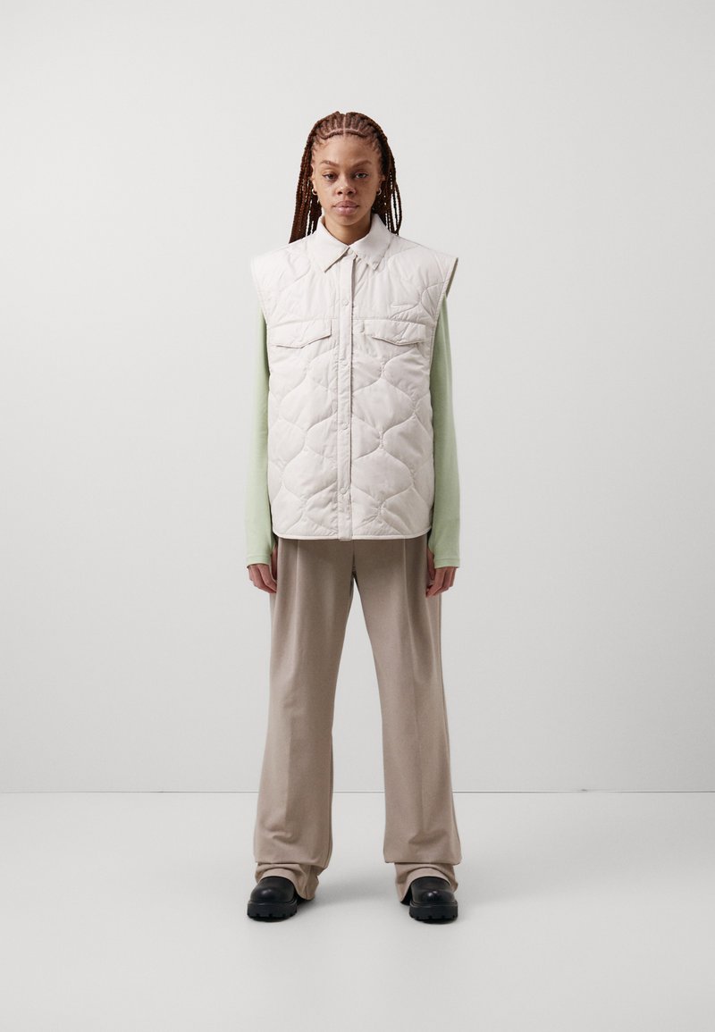 Άτομο που στέκεται φορώντας ένα αμάνικο επενδεδυμένο γκριζωπό γιλέκο, ανοιχτό πράσινο μακρυμάνικο μπλουζάκι, μπεζ φαρδιά παντελόνια και μαύρες ογκώδεις παπούτσια.