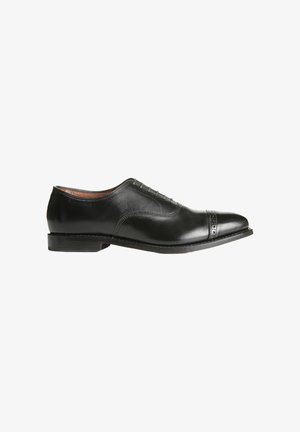 Black leather dress shoe with a smooth finish, cap toe design, perforated detailing, and a low heel. Features laces and a rounded toe.