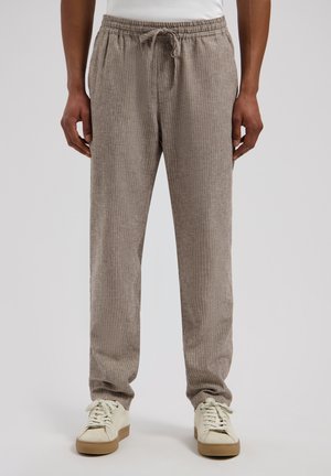 Man draagt beige geribbelde broek met trekkoord en gebroken-witte sneakers, staande tegen een effen achtergrond.