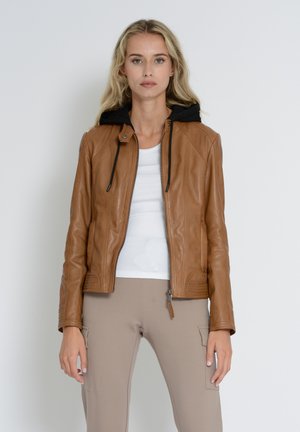 Veste en cuir beige avec capuche noire, fermeture éclair, poignets côtelés et poches latérales. Portée par-dessus un haut blanc et un pantalon beige.