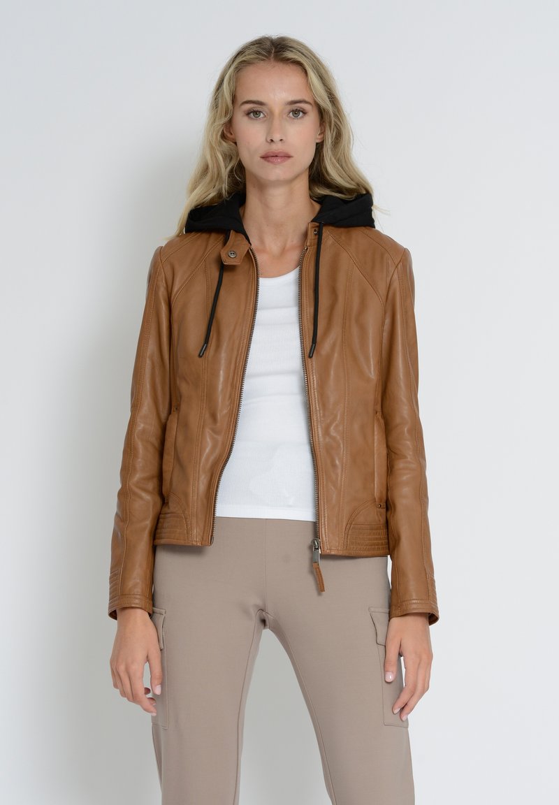 Veste en cuir beige avec capuche noire, fermeture éclair, poignets côtelés et poches latérales. Portée par-dessus un haut blanc et un pantalon beige.