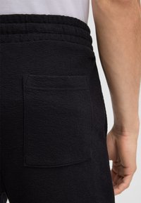 Zwarte gestructureerde joggers met een geribde tailleband en een patchzak op de achterkant, die een gebreide stofdetail benadrukken.