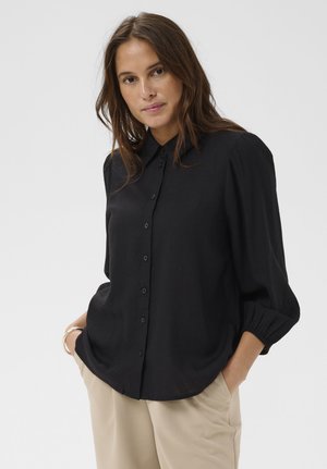 Mujer con cabello largo y marrón, vistiendo una blusa negra abotonada con mangas tres cuartos y pantalones beige, con las manos en los bolsillos, fondo neutro.