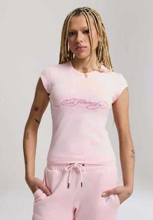 Jeune personne aux tresses blondes portant un t-shirt Ed Hardy rose clair et un pantalon de survêtement assorti, debout les mains dans les poches.