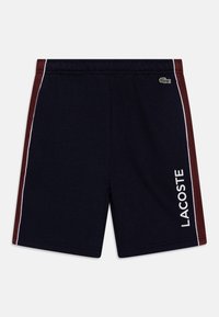 Tengerészkék rövidnadrág bordó oldalsó panelekkel, fehér díszítéssel és fehér "LACOSTE" logóval. Puha anyagból készült, elasztikus derékkal.