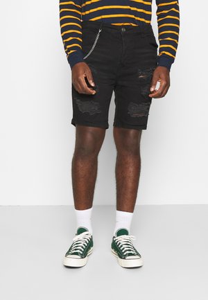 Black denim shorts with distressed details and rolled hems, featuring a chain accent. Paired with green canvas sneakers and white socks.
