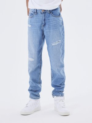 Jeans desgastados de color azul claro con un corte relajado, que presentan detalles rasgados en la parte frontal y un cierre de botón. Combinados con zapatillas blancas.