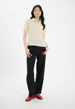 Polo shirt beige clair à manches courtes avec un tissage texturé, associé à un pantalon large noir et des mocassins rouges. Design simple et classique.