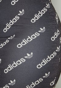 Leggings noirs avec une finition brillante et texturée, présentant un motif diagonal répétant le texte blanc "adidas" et le logo.
