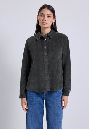Jeune femme portant des boucles d'oreilles en argent, une chemise boutonnée en velours côtelé gris foncé et un jean bleu, sur fond clair uni.
