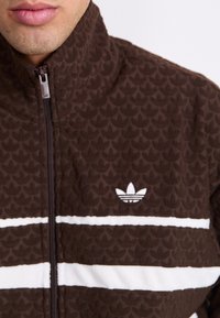 Ruskea vetoketjullinen takki, jossa on teksturoitu kuvio, valkoisia vaakasuoria raitoja ja brodeerattu logo rinnassa. Pehmeä, fleecen kaltainen materiaali.