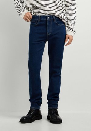 Person trägt dunkelblaue Jeans mit geradem Bein, schwarze Schnürstiefel und ein weißes Langarmshirt mit schwarzen horizontalen Streifen.