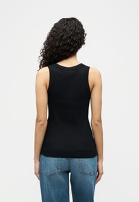 Femeie cu păr lung și creț, purtând un top negru fără mâneci și blugi albaștri, privind cu spatele spre fundalul alb simplu.