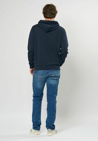Sudadera navy con corte relajado, que incluye un bolsillo canguro y capucha con cordón. Combinada con jeans azules y zapatillas de color claro.
