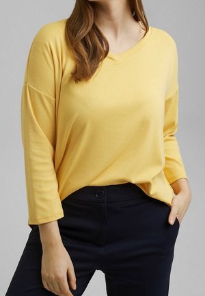 Kobieta ubrana w żółtą bluzkę z długim rękawem i dekoltem w serek oraz czarne spodnie, z jedną ręką wsuniętą do kieszeni, na jednolitym szarym tle.