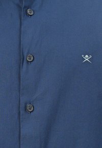 Chemise boutonnée bleu marine en tissu texturé avec six boutons foncés. Présente un petit design brodé en bleu clair sur la poitrine.