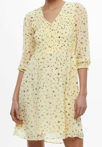 Keltainen kukkamekko, jossa on V-pääntie, rypytetty yläosa ja kolmanneksen mittaiset puffihihat. Kevyt, läpikuultava kangas, jossa on pieniä monivärisiä kukkakuoseja.