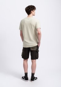Lichtbeige katoenen T-shirt met korte mouwen, gecombineerd met donkere cargoshorts met zijzakken en contrasterende stofpatches. Zwarte sneakers.
